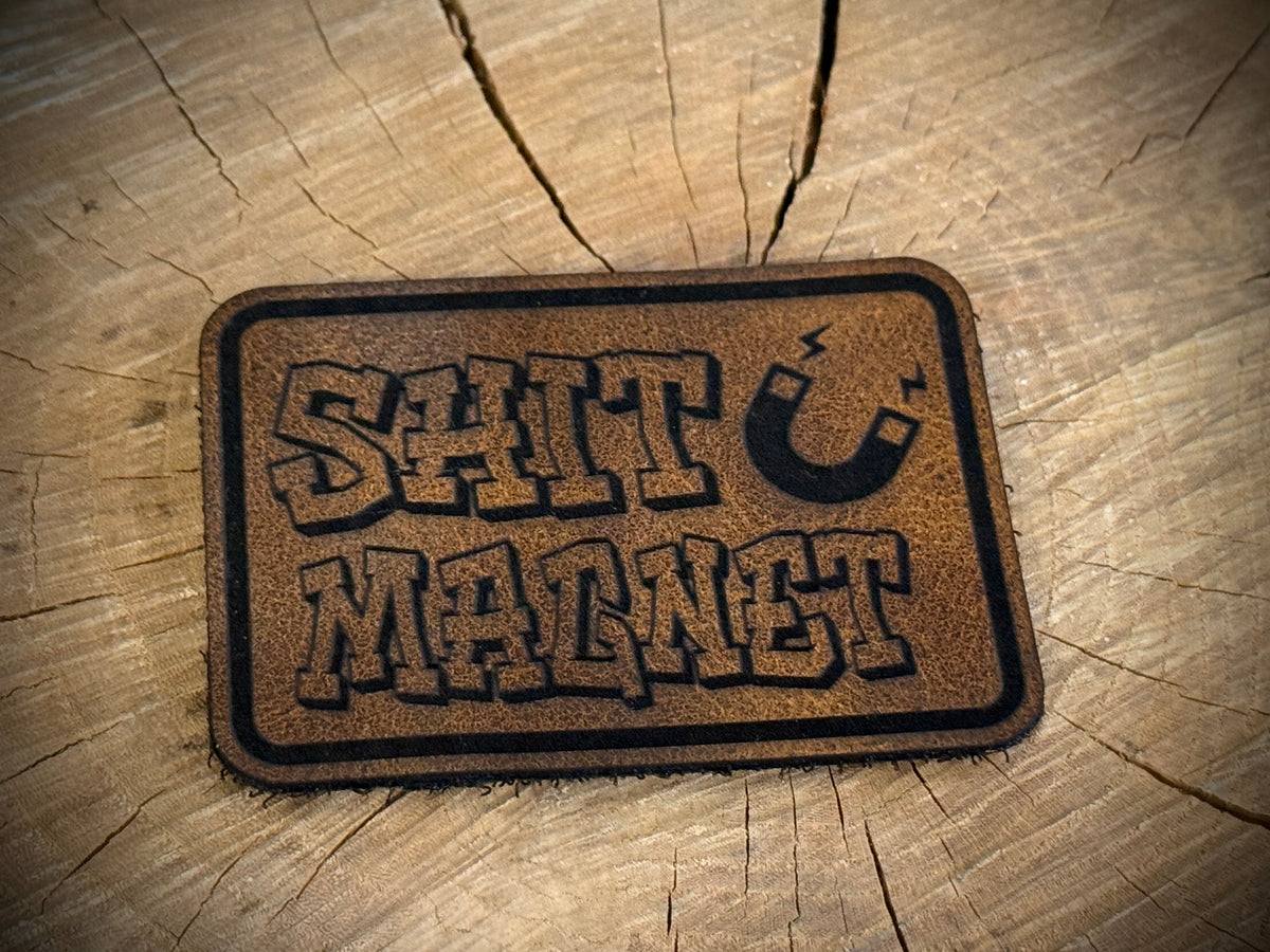 Leather velcro backed Morale Patch with 'Shit Magnet' text on a wooden surface