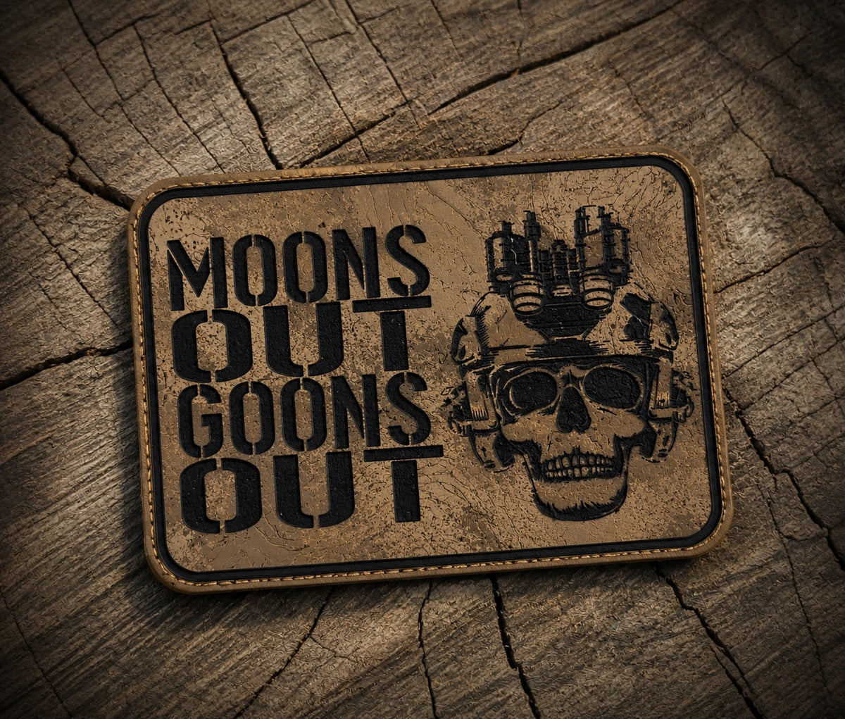 A rectangular tan tactical patch with rounded corners, featuring the text 'MOONS OUT GOONS OUT' in black stencil font alongside a black skull wearing a helmet with mounted lights, placed on a textured wood background.