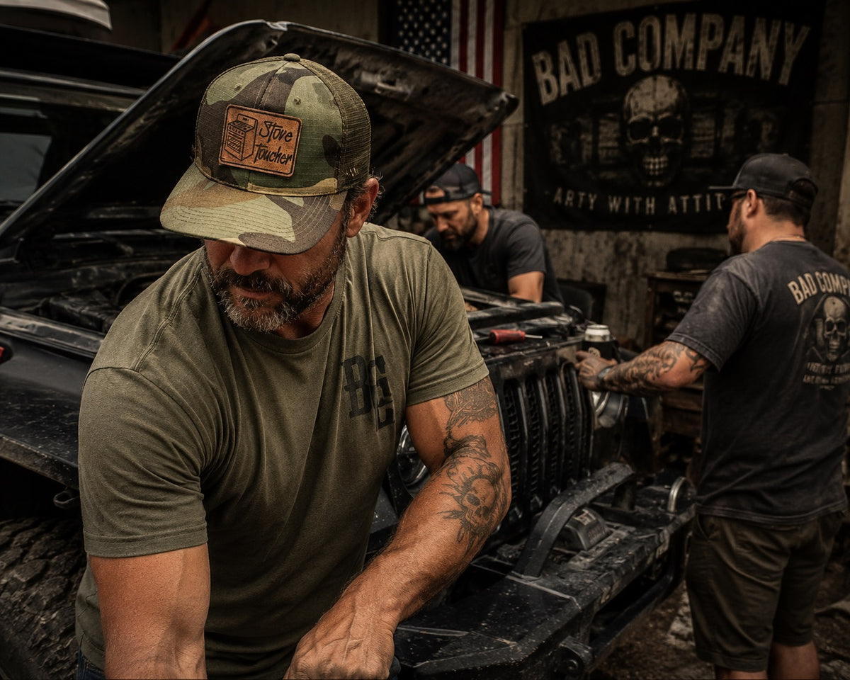 A man in a camouflage truck cap WITH A  FUNNY MORALE PATCH THAT SAYS "STOVE TOUCHER" and olive green t-shirt works on a vehicle in a garage, with another man in a black 'Bad Company' t-shirt and another man in the background near an American flag and a 'Bad Company' banner.